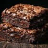 Fudgy sweet potato brownies stacked on dark parchment paper with melted chocolate chips and flaky sea salt