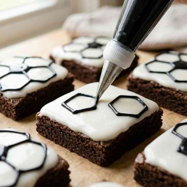 Decorating soccer brownies with black gel icing piped in a soccer ball pentagon pattern on white vanilla icing
