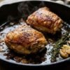 Pan seared chicken thighs in a cast-iron skillet with crispy golden brown skin, fresh thyme, and smashed garlic cloves